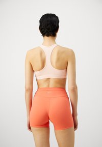 Sujetador deportivo rosa claro con diseño de espalda cruzada y tirantes elegantes, combinado con pantalones cortos ajustados naranjas que presentan un sutil logo de Nike en la cinturilla.