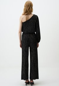 Zwart one-shoulder jumpsuit met wijde pijpen, textuurstof met metallic draden, verstelbare taille en lange mouwen. Hiel-schoenen zichtbaar.