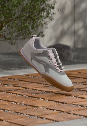 Baskets Calvin Klein gris clair et blanc avec des lacets roses flottant au-dessus d’un sol extérieur en carreaux de bois avec une plante verte en arrière-plan.