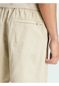 Pantaloni corte beige con vita elastica, dotati di una tasca posteriore con zip e una trama di tessuto liscia. Tasche laterali per le mani visibili.