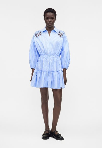 Γυναίκα που φοράει ένα γαλάζιο ριγέ φόρεμα πουκάμισο με λεπτομέρειες κοπής στους ώμους και μαύρα παπούτσια Mary Jane, στέκεται μπροστά σε λευκό φόντο.