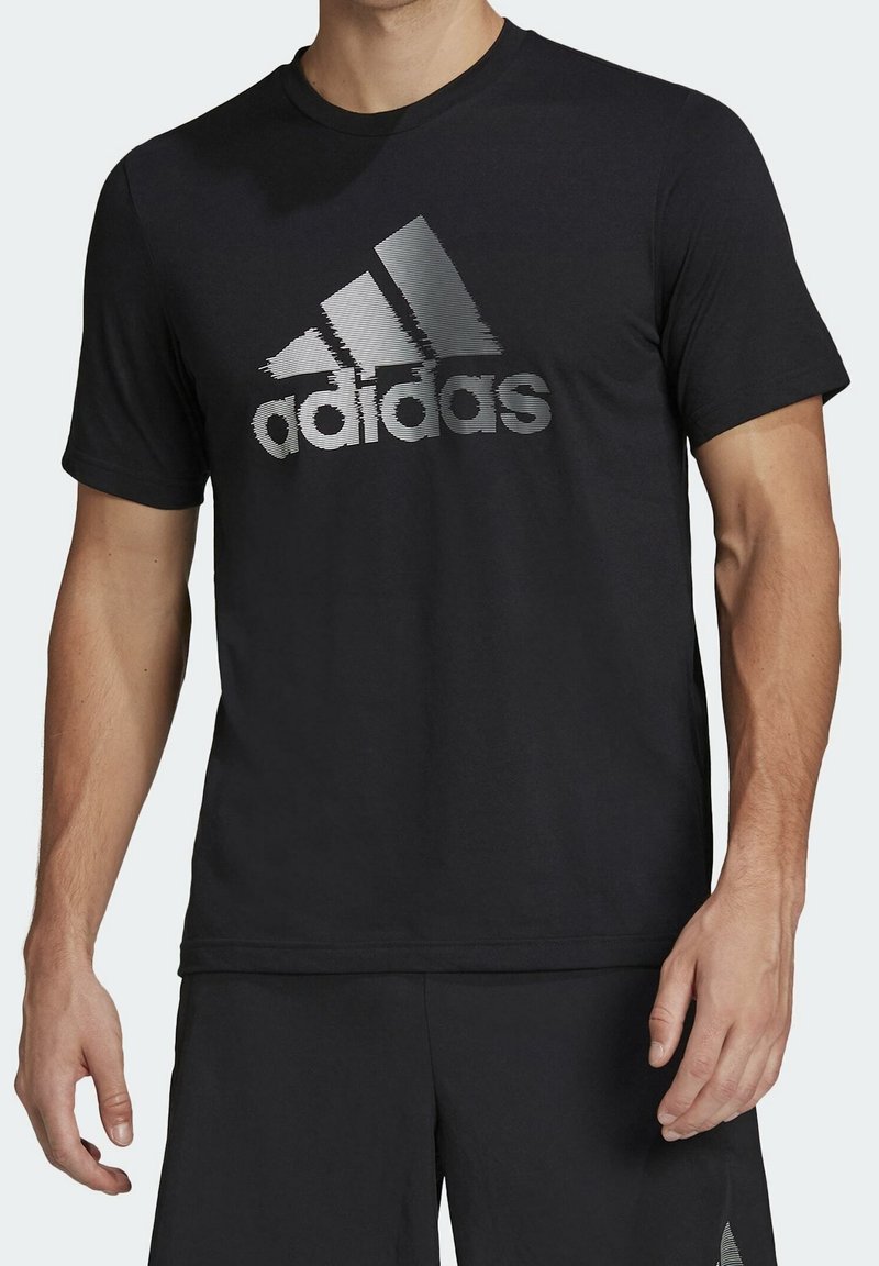 Mies, joka on pukeutunut mustaan Adidas-paitaan, jossa on suuri hopeinen Adidas-logo rinnassa, ja mustiin shortsisiin, valkoista taustaa vasten.