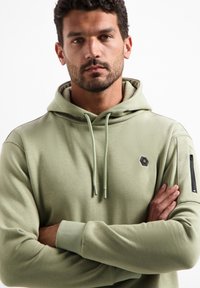 Lichtgroene capuchontrui met een voorzak, geribbelde manchetten en een ritszak op de mouw. Voorzien van een zeskantig logo.