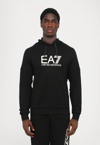 Felpa nera con cappuccio caratterizzata da un grande logo bianco EA7 Emporio Armani sulla parte anteriore. Realizzata in morbido tessuto con tasca a marsupio.