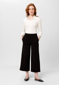 Blusa bianca a maniche lunghe con bottoni, abbinata a pantaloni neri a vita alta e a gamba larga. Il modello indossa delle scarpe slingback nere. Sfondo neutro.