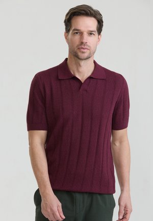 Homme aux cheveux bruns courts portant un polo bordeaux texturé à manches courtes et un pantalon vert foncé, debout devant un fond uni.