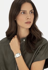 Silberne und goldene Metalluhr mit einem quadratischen weißen Ziffernblatt, am Handgelenk getragen. Die Person hat lange dunkle Haare und trägt eine dunkle Lederjacke.