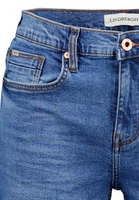 Nærbillede af blå denimjeans i taljen med frontlomme, lille møntlomme, kobbernitter, bæltestrop og Lindbergh-mærkat inden i linningen.