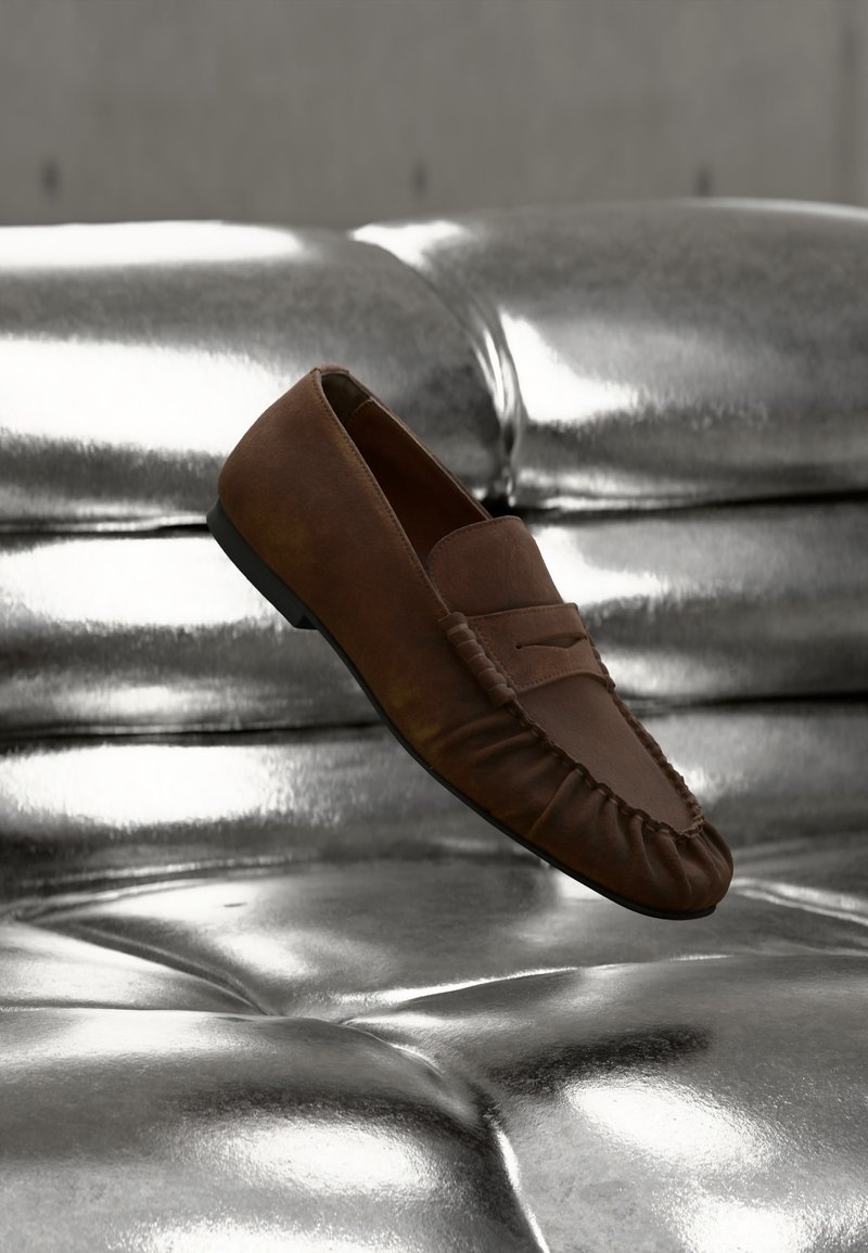 Καφέ δερμάτινο μοκασίνι παπούτσι που εμφανίζεται πάνω από μια γυαλιστερή, μεταλλική ασημένια επιφάνεια ή καναπέ με κουμπιά.