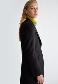 Cappotto lungo nero con due bottoni e design elegante, indossato sopra un dolcevita verde a coste. Presenta una texture liscia e una vestibilità su misura.