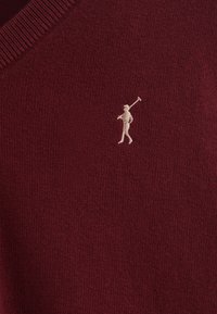 Bordeauxstickad tröja med V-hals, med en liten beige broderad figur som håller en golfklubba på övre vänstra sidan.