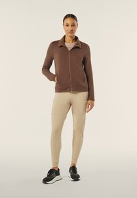 Giacca marrone con zip e colletto testurizzato, abbinata a pantaloni beige a gamba affusolata e sneakers nere con suola bianca.
