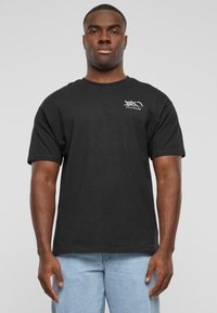 Sort bomulds t-shirt med korte ærmer, rund halsudskæring og hvidt trykt logo på venstre bryst. Båret med lyseblå jeans.