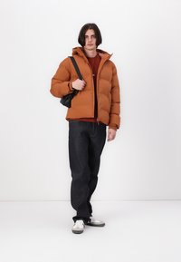 Brun PU-jakke med hætte, lynlås og elastiske manchetter. Båret over en rød sweater med logo, parret med mørke jeans og sneakers.