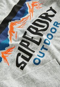 Grijze stoffen T-shirt met het "Superdry" logo in zwart en blauw, met oranje geborduurde accenten van gestileerde bergen.