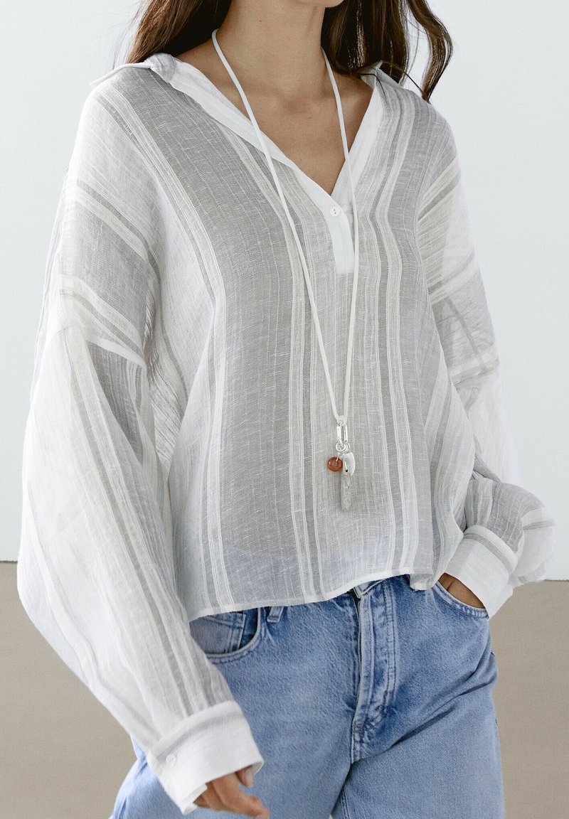 Femme portant un chemisier blanc rayé semi-transparent ample avec un décolleté en V profond sur un jean bleu, accessoirisé d'un collier pendentif.