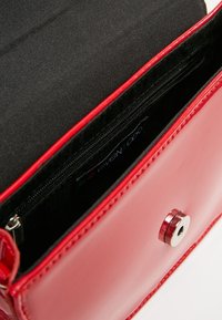 Bolso de mano de charol rojo con una textura suave. El interior cuenta con un forro de tela negra y un bolsillo con cremallera. Cierre de hardware en plata.