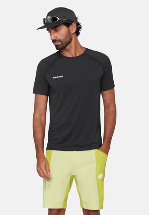 Mand iført sort Mammut t-shirt, gul-grønne shorts, sort kasket med solbriller, står med den ene hånd i lommen og kigger ned.