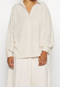 Krämfärgad skjorta med knapptryckning och krage, långa puffärmar och elastiska manchetter, tillverkad av ett mjukt, texturerat tyg. Enkel design.