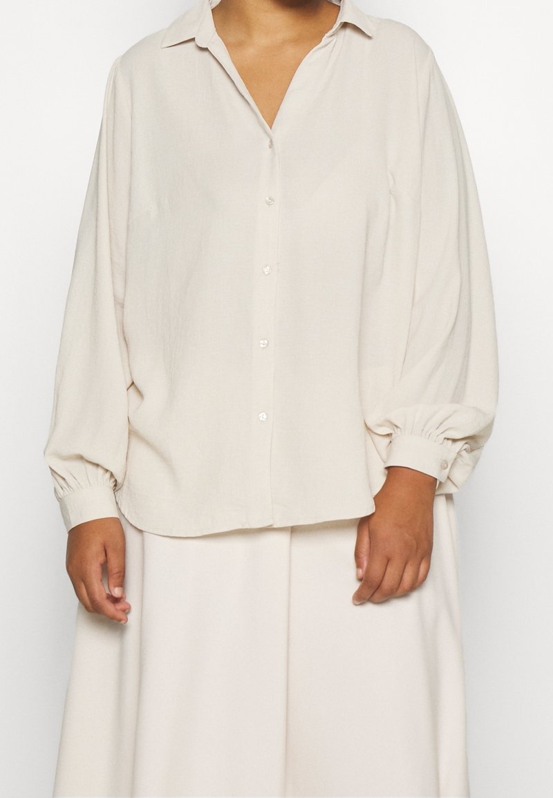 Krämfärgad skjorta med knapptryckning och krage, långa puffärmar och elastiska manchetter, tillverkad av ett mjukt, texturerat tyg. Enkel design.