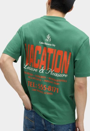 Persona che indossa una maglietta verde con grande scritta arancione "VACATION" e informazioni sul servizio di svago di Lake Havasu City sul retro.
