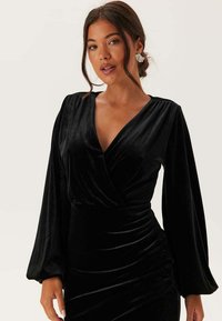 Sort velour kjole med V-hals, lange pufærmer og rynk ved taljen for en tætsiddende elegance. Glat tekstur med subtil glans.