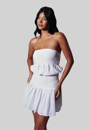 Kvinde med langt sort hår iført en stropfri hvid øjlet kjole med flæsedetaljer, stående foran en ensfarvet grå baggrund.