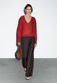 Maglione rosso lavorato a maglia con scollatura a V profonda e vestibilità ampia, abbinato a pantaloni larghi marroni. Il modello tiene in mano una borsa rotonda marrone dalla superficie texture.