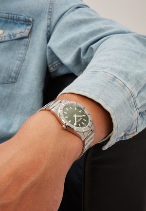 Sølv rustfrit stål ur med en mørkegrøn urskive, hvide timemarkører og en poleret finish. Båret på et håndled med en blå denimjakke.