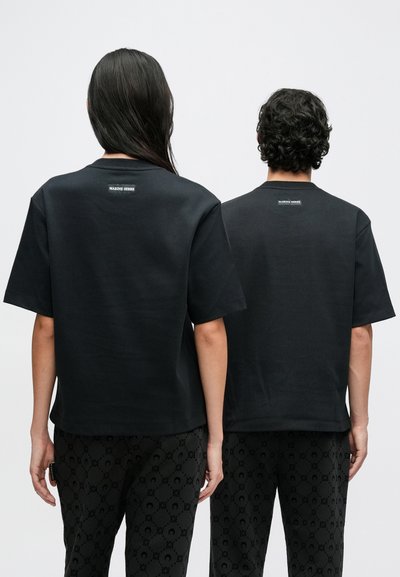 T-shirts de algodão pretas com mangas curtas, apresentando uma pequena etiqueta de logo nas costas. Usadas com calças pretas estampadas.