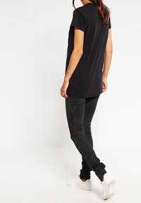 Schwarzes Baumwoll-T-Shirt mit kurzen Ärmeln, längerem Rückensaum, kombiniert mit dunklen Slim-Fit-Jeans und weißen Sneakers. Glatte Textur, lässiger Schnitt.