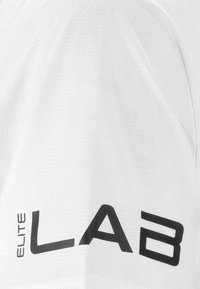 Haut de sport blanc en tissu texturé, arborant un logo « ELITE LAB » noir sur le côté gauche. Design simple, sans motifs supplémentaires.