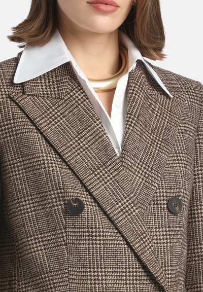 Brown and cream houndstooth double-breasted blazer with black buttons, paired with a white shirt and a gold circular necklace.