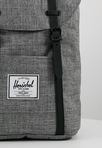Sac à dos en tissu gris texturé avec sangle noire à boucle et étiquette rectangulaire blanche de la marque Herschel sur la poche avant.
