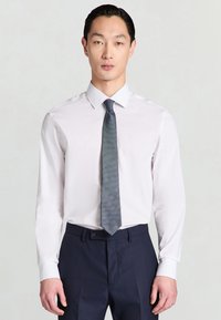 Weißes Hemd mit langen Ärmeln und einem Hemdkragen, kombiniert mit einer strukturierten dunkelblauen Krawatte. Dazu werden dunkelblaue Hosen getragen.