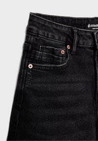 Gros plan sur un jean en denim noir montrant la poche avant avec des rivets en cuivre, une boucle de ceinture visible, des détails de couture et une étiquette de marque à l'intérieur de la ceinture.