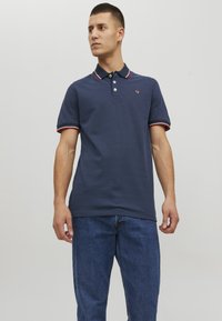 Mornarska polo majica z ovratnikom in manšetami v rdeče-beli črtasti vzorec, z manjšim logotipom na prsih. Kratke rokave in teksturirano blago.