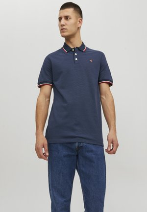 Camisa polo azul marino con cuello y puños a rayas rojas y blancas, con un pequeño logo en el pecho. Mangas cortas y tejido texturizado.