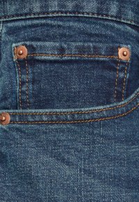 Jeans i denim med en bakficka som har kopparnitar och kontrasterande gyllene sömmar. Blått tyg med en texturerad yta och lätt blekning.