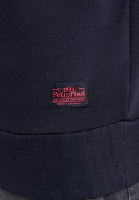 Námořnická modrá mikina s žebrovaným lemem. Obsahuje tkanou štítkovou etiketu v červené a černé barvě s nápisem "Petrol Ind 1989 Driven by Heritage."