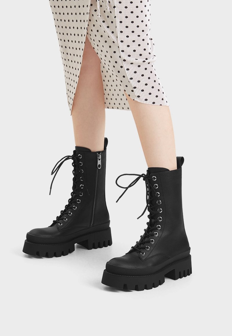Bottes de combat noires épaisses à lacets avec fermeture éclair latérale, portées avec une jupe blanche à pois noirs et une fente devant.