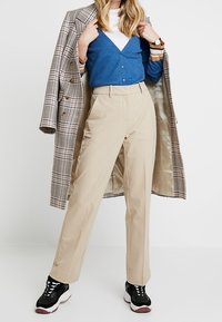 Beige högmidjade byxor med rak passform, kombinerat med en blå ribbad cardigan och en rutig kappa, kompletterat med svarta sneakers.