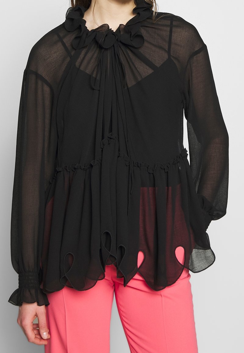Femme portant un chemisier noir transparent à manches longues avec des volants et un ourlet festonné sur un caraco noir, associé à un pantalon rose vif.