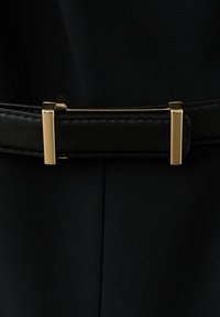 Ceinture en cuir noir avec une fine boucle rectangulaire dorée sur un fond en tissu sombre.