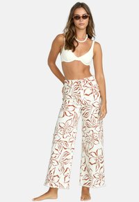 Weiße, weit geschnittene Blumenhosen mit roten Blumenmustern, kombiniert mit einem cremefarbenen Bikinioberteil. Das Modell trägt Sonnenbrillen, ist barfuß und trägt eine Halskette.