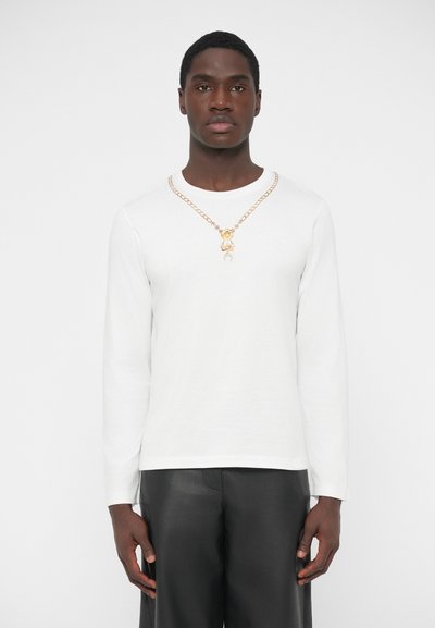 Chemise blanche à manches longues avec un col rond, associée à un pantalon en cuir noir. Un collier chaîne en or présente un pendentif en forme d'ours.