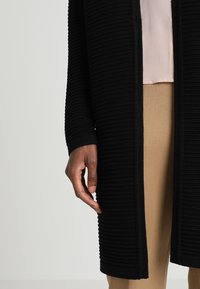 Svart ribbad cardigan tillverkad av texturerad tyg, med långa ärmar och öppen front. En beige topp sticker fram under.