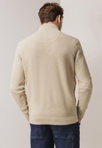 Beige geribbelde trui met een hoge kraag en lange mouwen, voorzien van een gestructureerde afwerking en een aansluitende onderzoom, gedragen met donkere jeans.