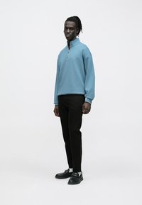 Sudadera azul claro con cremallera y corte holgado, combinada con pantalones negros y zapatos negros. Tejido liso y diseño minimalista.