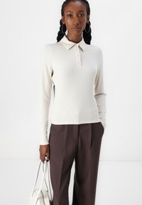 Ribbestrikket langermet polo-skjorte i off-white, med krage, tre knapper, og en figursydd silhuett, paret med mørke bukser.